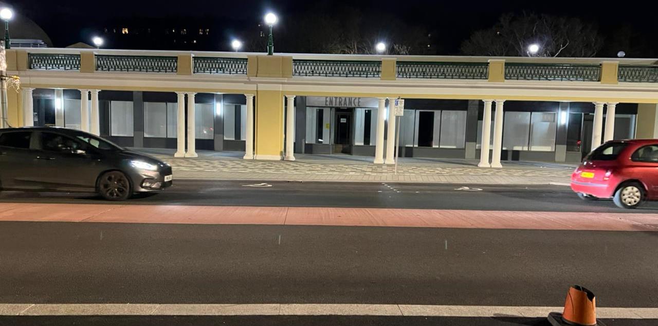A night-time photograph of a commercial building featuring a colonnade and balcony, with cars parked on the street in front.