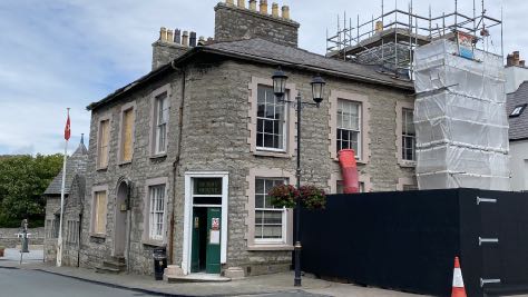 A photograph of a two-story stone building undergoing renovation, featuring scaffolding and white sheeting on the right side and a black construction hoarding in the foreground.