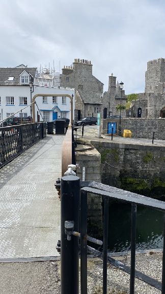 A street-level photograph showing a bridge with black railings leading towards historic stone buildings and a castle-like structure in the background.