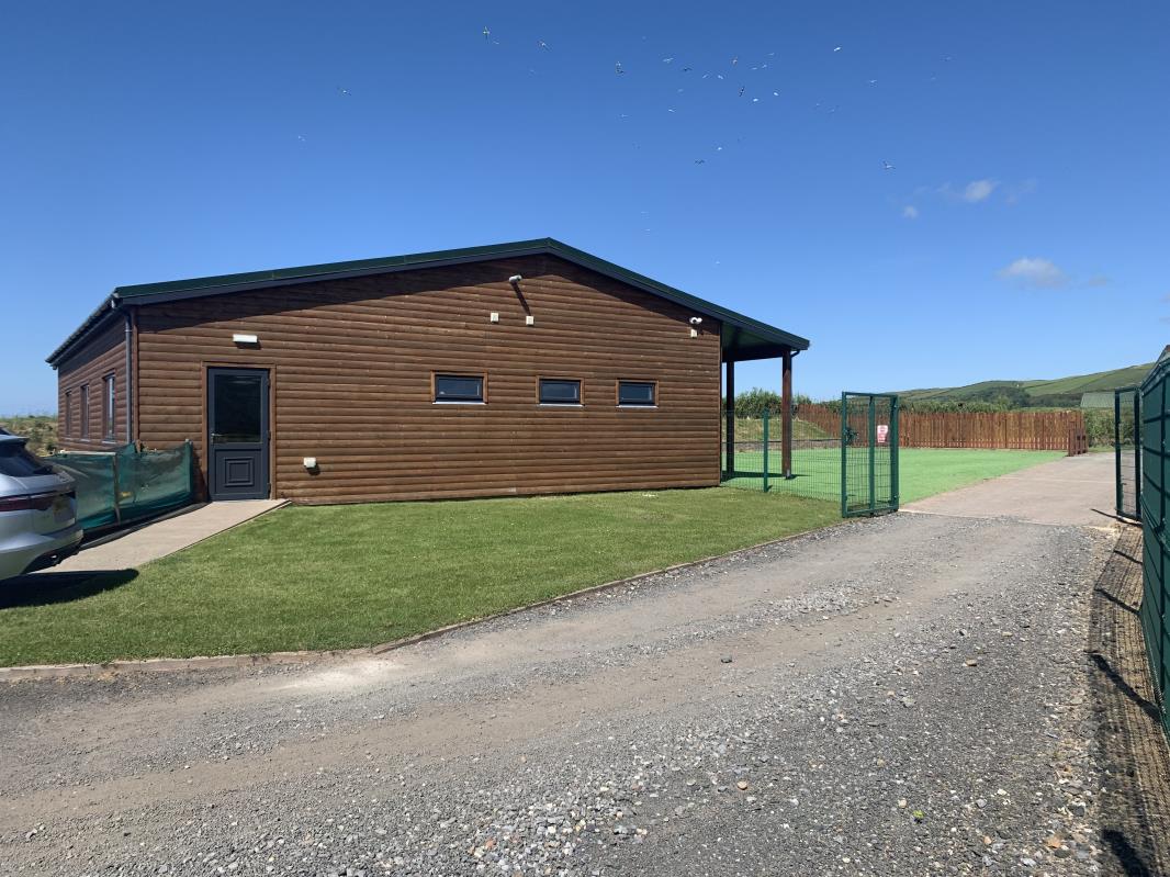 A single-story building with brown horizontal cladding and a covered porch area is shown with a gravel driveway in the foreground. A green fenced enclosure is visible to the right in a rural setting.
