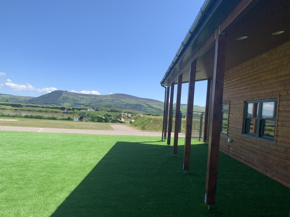 A photograph showing the exterior of a wooden-clad building with a covered porch and artificial turf lawn. The background features a scenic view of a lake and rolling hills under a blue sky.