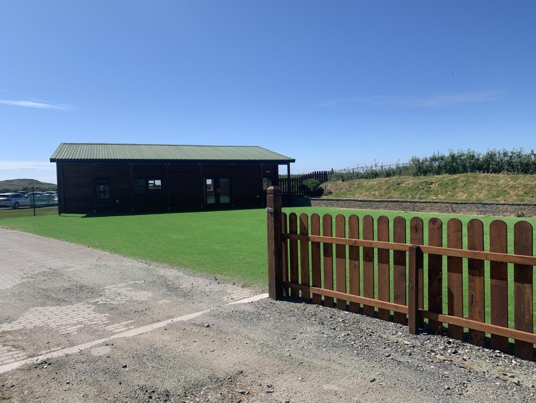 A photograph showing a single-story wooden building with a green roof situated next to a wooden fence and a patch of green lawn.