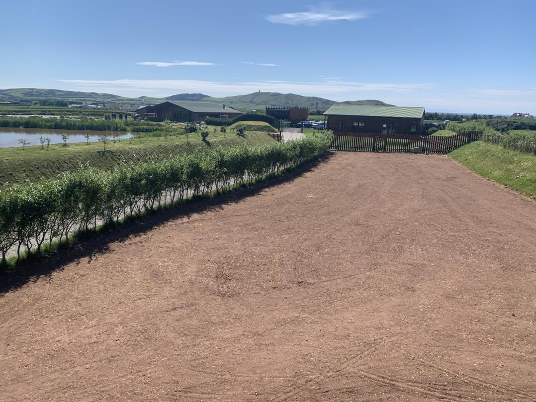 A sunny outdoor photograph showing a gravel driveway leading towards a small wooden outbuilding and larger agricultural structures near a body of water with coastal views in the distance.