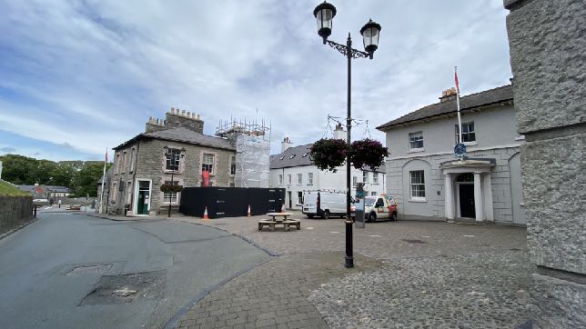 A street-level photograph of a town square in Castletown showing stone buildings, one of which is under renovation with scaffolding and hoarding.