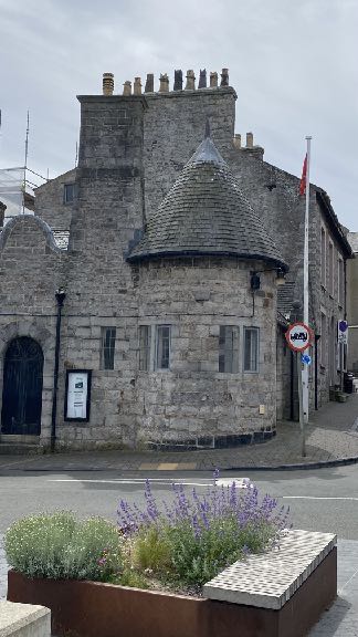 A photograph of a historic stone building with a turret and conical roof, showing scaffolding on the left side and a flag pole on the right.