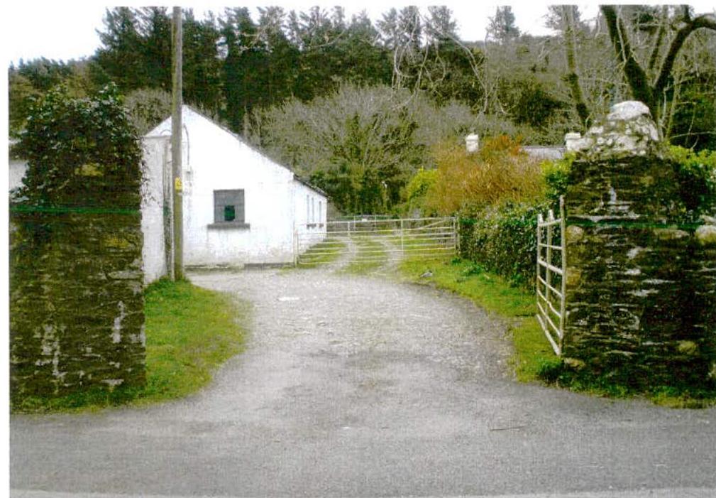 A photograph showing a white single-story building set back from a road, accessed via a gravel driveway with stone gateposts and metal gates. The scene is rural with trees in the background.