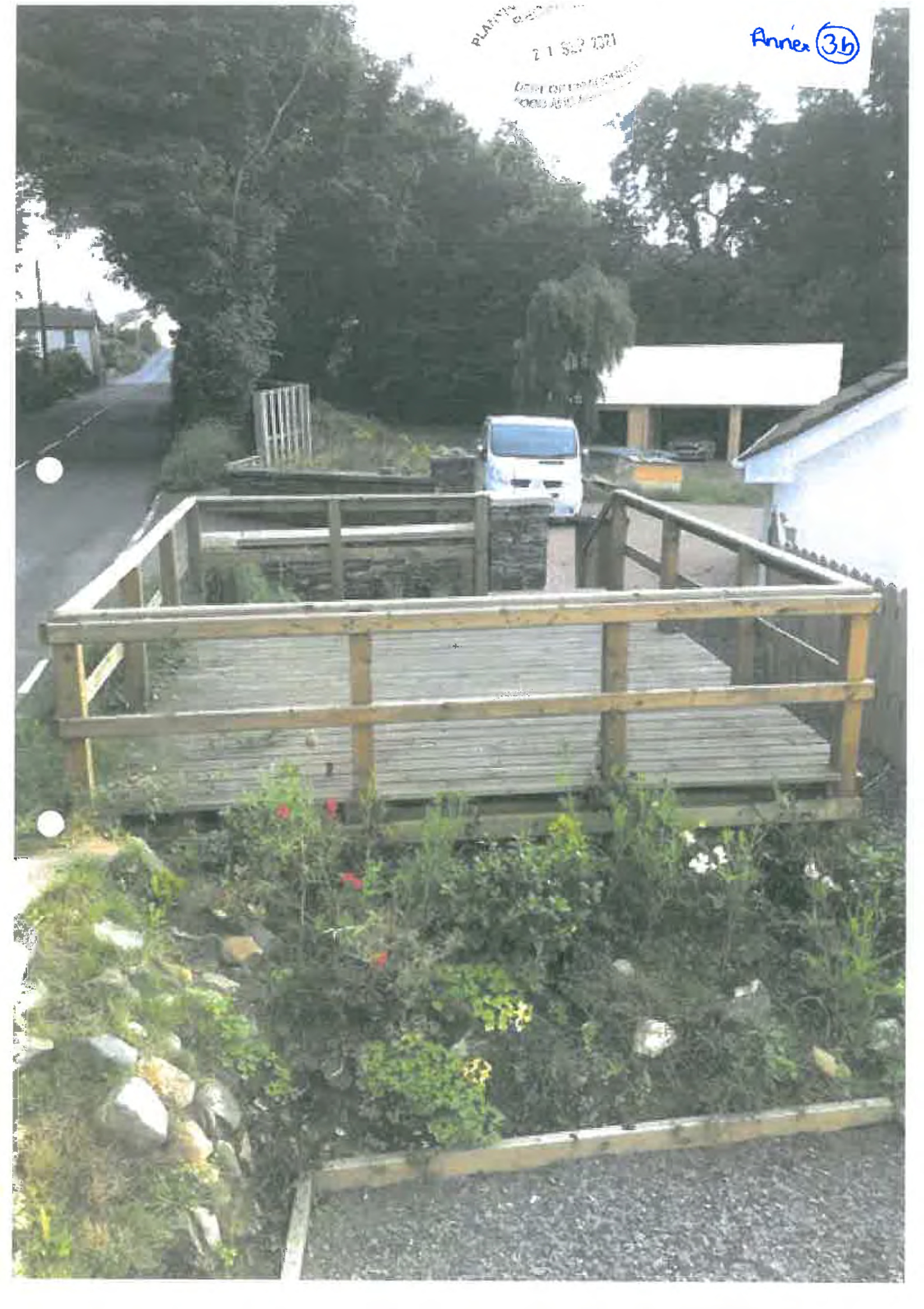 A photograph showing a newly constructed wooden raised deck with railings situated in a garden area next to a road.