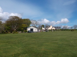 A landscape photograph showing a large grassy field in the foreground with existing white buildings and trees in the background under a blue sky.