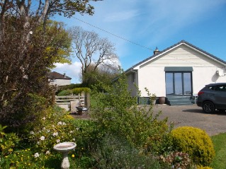 A photograph showing an existing white bungalow with a driveway, garden, and parked car in a rural setting.