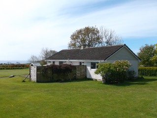 A photograph of a single-story white building with a dark roof situated in a grassy rural field, likely the existing property on the site.