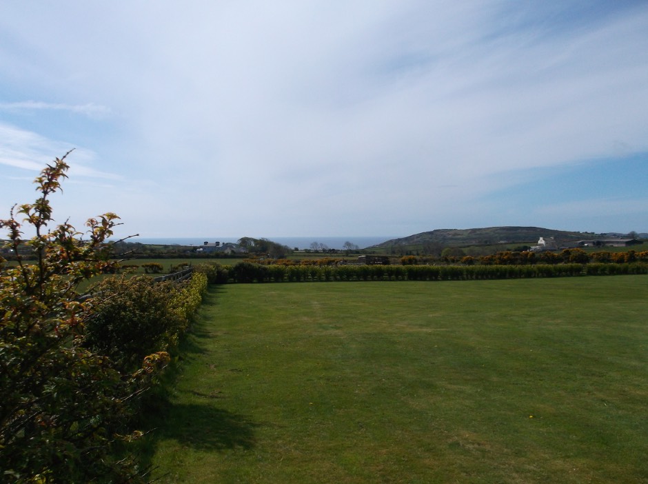 A landscape photograph showing a large grassy field bordered by a hedge, with a view of the sea and distant hills in the background.