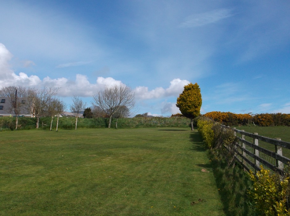 A photograph showing a grassy field with a wooden fence running along the right side and trees in the background, likely the site for the proposed development.