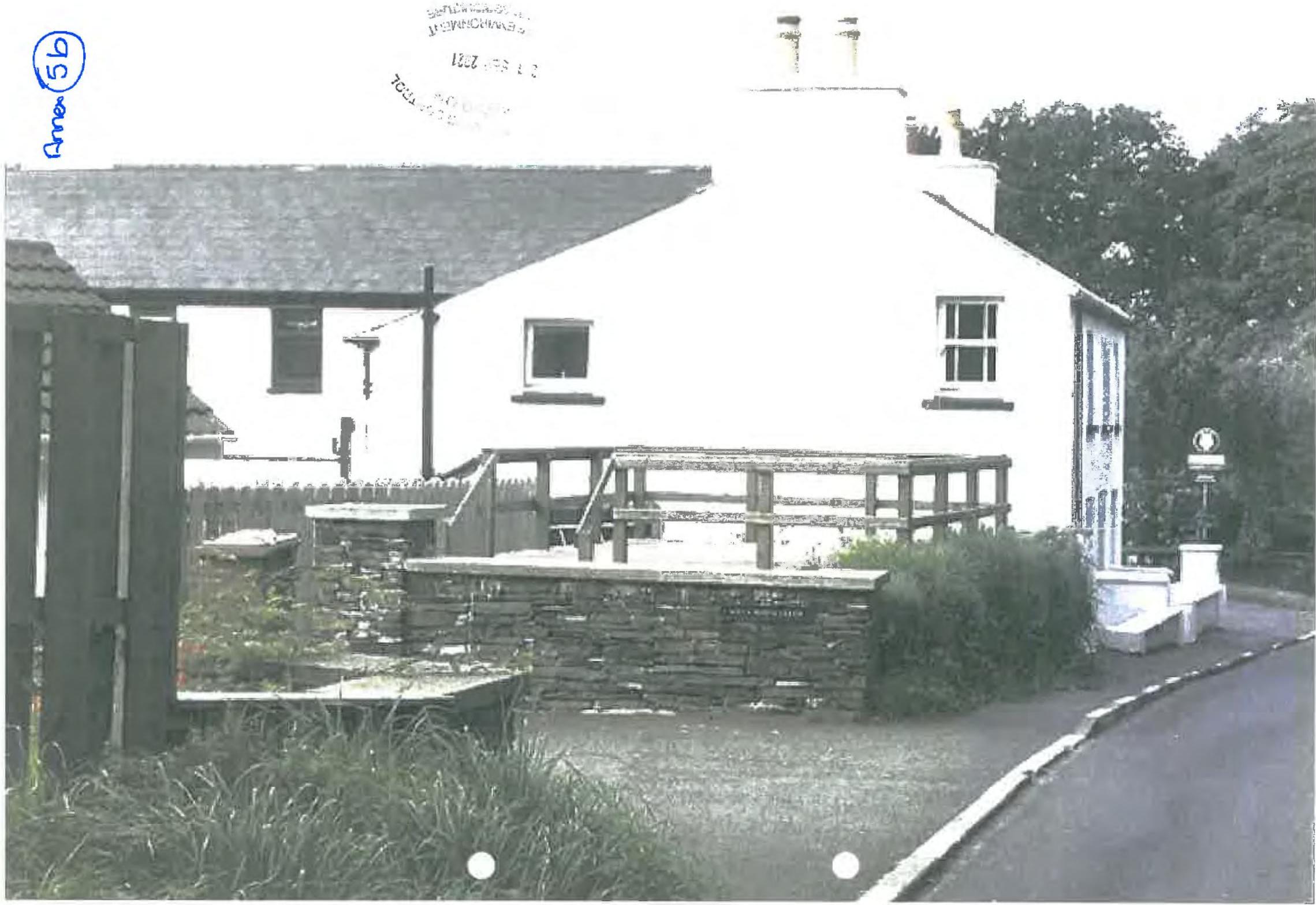 A black and white photograph showing a white cottage with a wooden deck and stone retaining walls at the rear, situated next to a road.