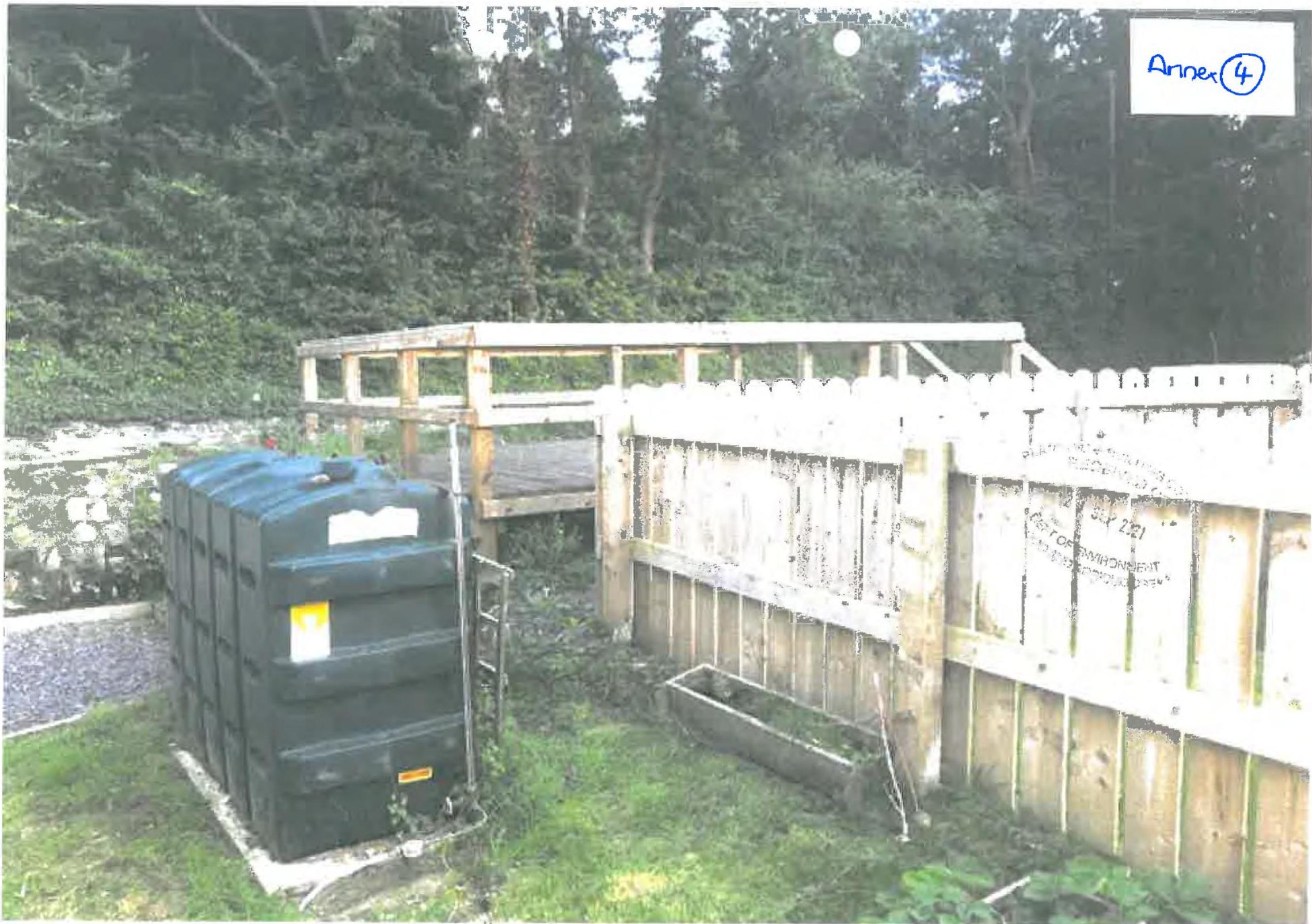 A photograph showing a wooden raised deck structure with railings situated next to a wooden fence and a large green water tank in a garden area.