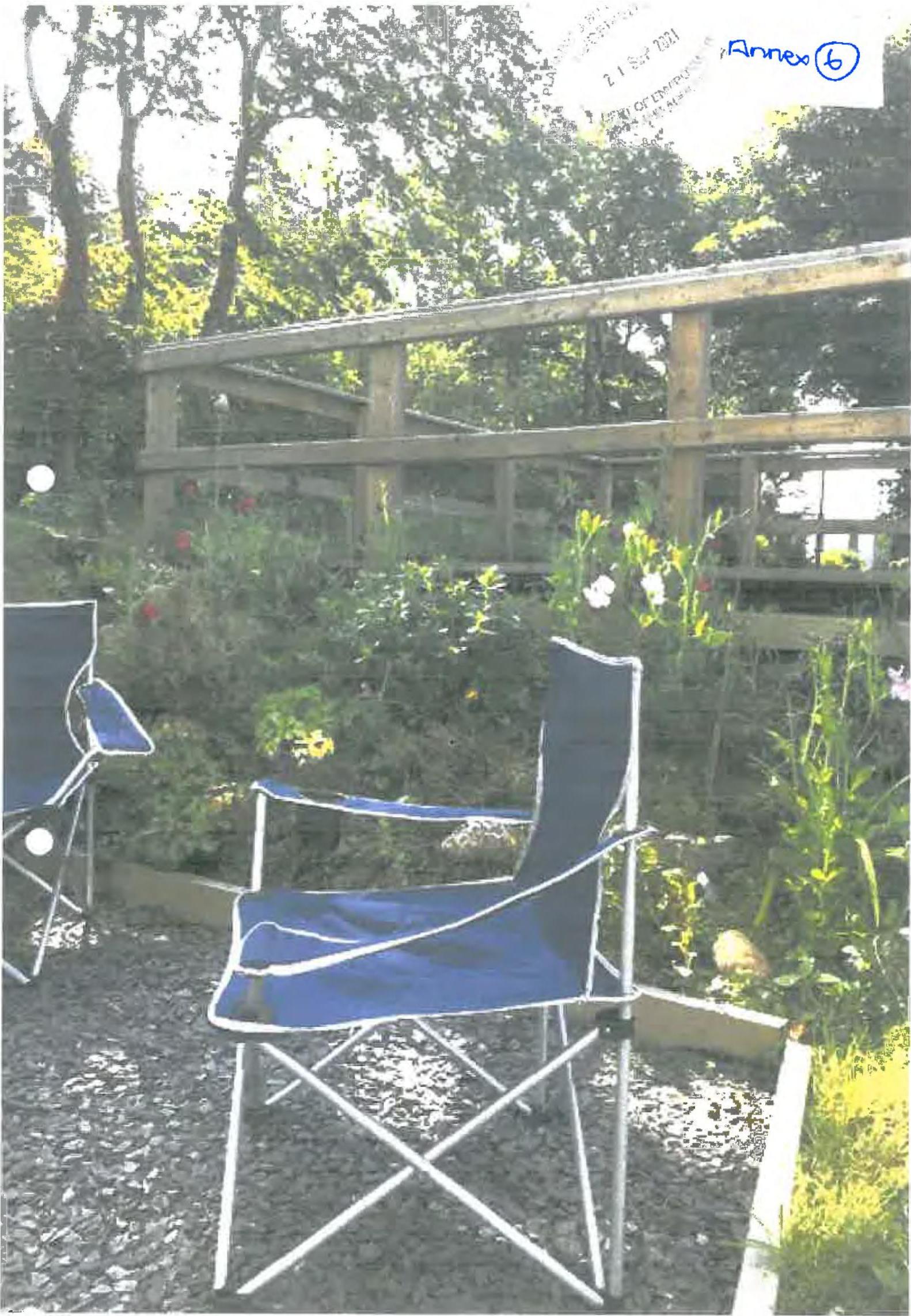 A photograph showing a garden area with blue camping chairs on gravel, a wooden fence or railing structure, and surrounding vegetation.