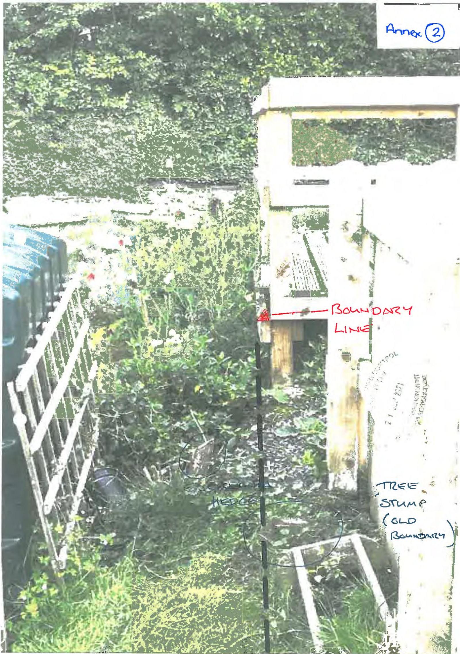 A photograph of an outdoor area showing a wooden fence or gate structure next to a metal fence, annotated with handwritten notes indicating a boundary line and tree stump.