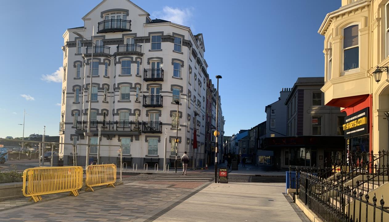 A street-level photograph showing a large white multi-story building with balconies, likely the proposed development, situated on a paved promenade with yellow barriers in the foreground.