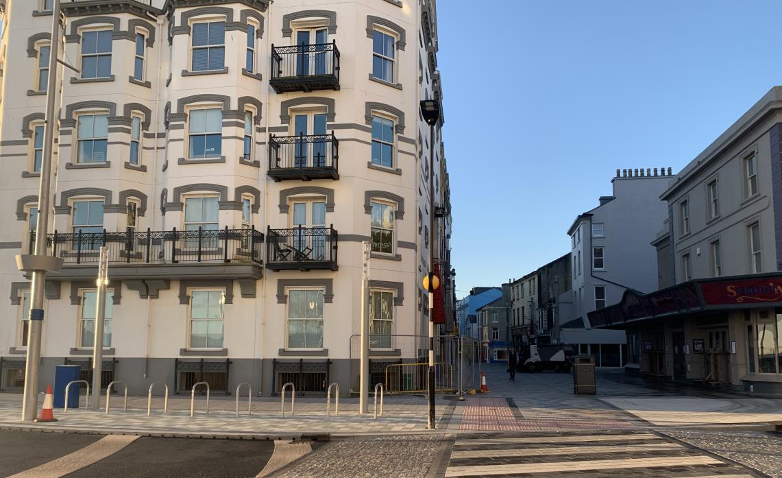 A street-level photograph showing a large white multi-story building with balconies on the left and a paved street scene with other buildings on the right.