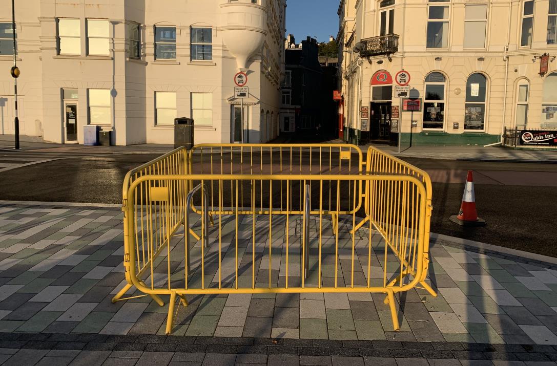 A street-level photograph showing yellow crowd control barriers and a traffic cone on a paved sidewalk in front of commercial buildings.