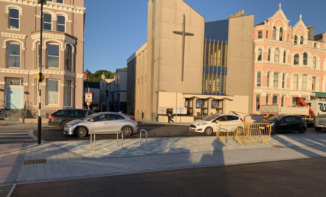 A street-level photograph showing a modern concrete building with a large cross on its facade, flanked by traditional Victorian-style buildings in a town center. The foreground features a paved area with parked cars,...