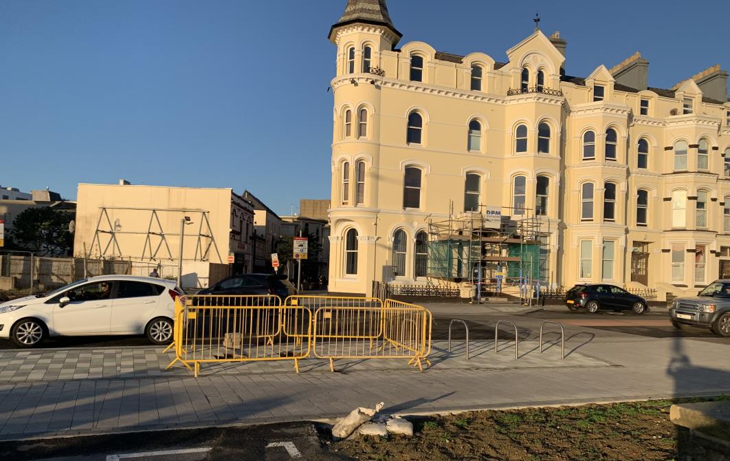 A street-level photograph showing a large, cream-colored, multi-story building with scaffolding on the ground floor, likely undergoing renovation or construction. The foreground features a paved area with yellow barri...