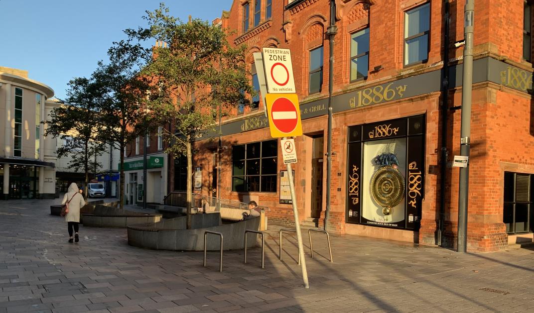 A street-level photograph showing a red brick commercial building with '1886' signage and a pedestrian zone sign in the foreground. The scene includes paved landscaping, benches, and trees along the sidewalk.