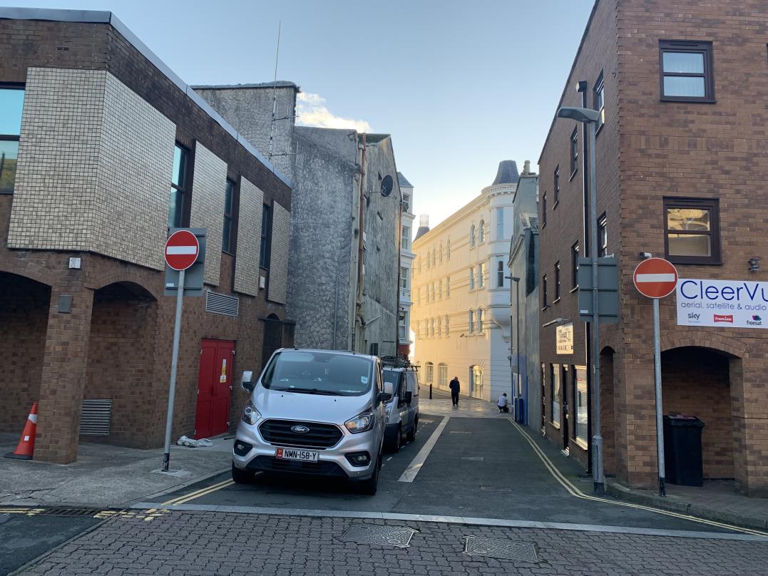 A street-level photograph of a narrow urban lane in Douglas, featuring brick buildings on either side, a parked silver van, and 'No Entry' traffic signs.