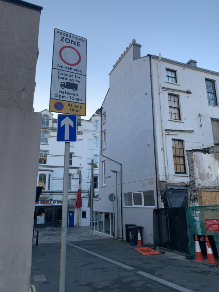 A street-level photograph showing a white building undergoing renovation with black hoarding and traffic cones, situated next to a tall pedestrian zone signpost.