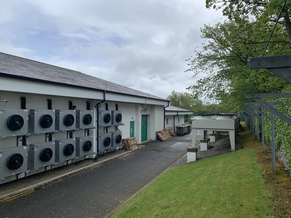 This photograph shows the exterior elevation of a single-story commercial building featuring a large array of industrial cooling units mounted on the wall. The view includes a paved access area, waste bins, and an adj...