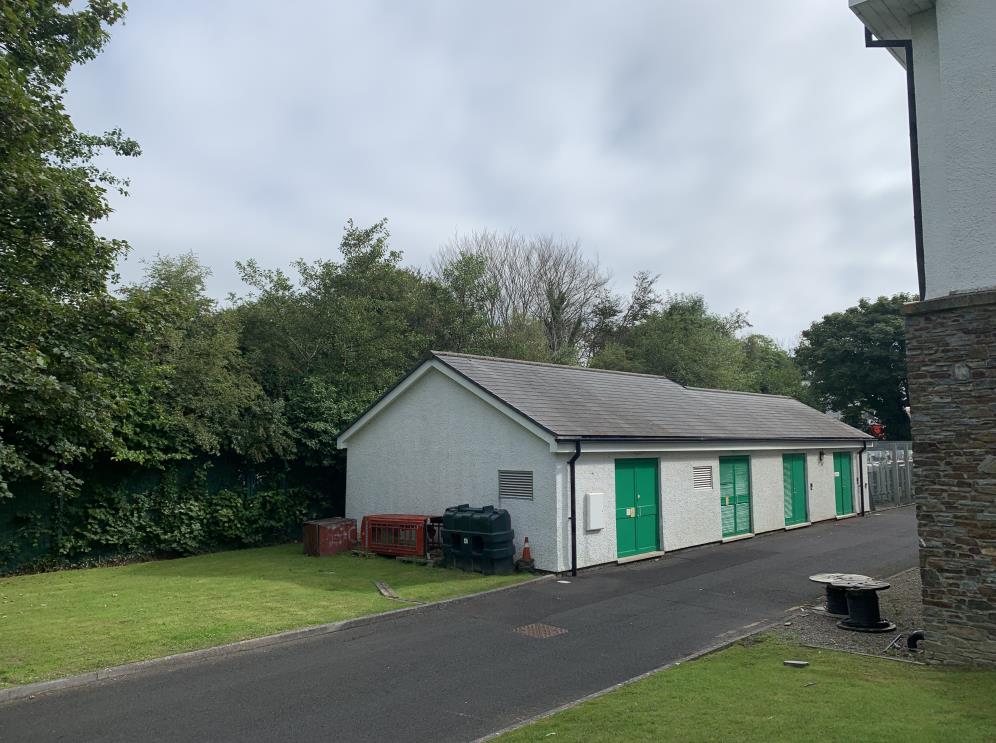 A photograph showing a long, single-story white building with a grey tiled roof and multiple green doors, situated next to a paved area and trees.