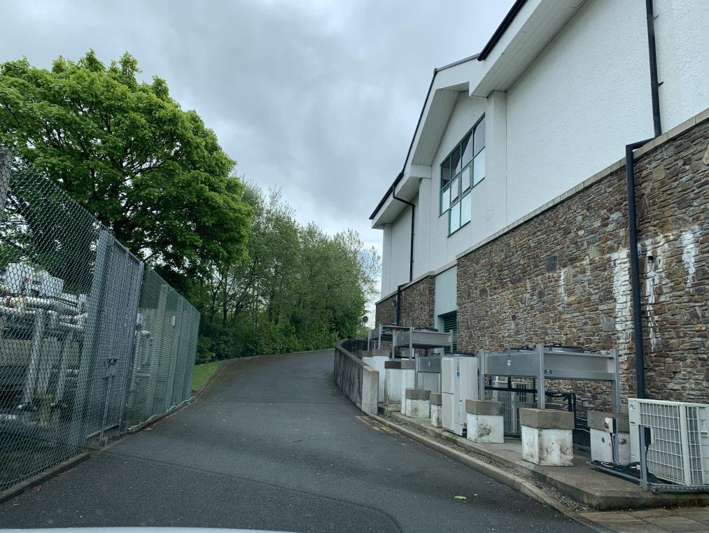 A photograph showing the side elevation of a white and stone building with multiple cooling units installed along the base. A paved path runs alongside the units, bordered by a chain-link fence on the left.