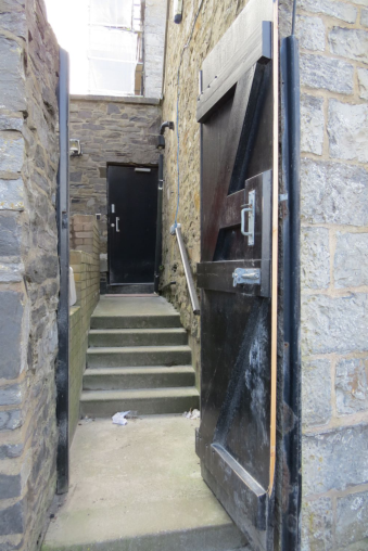 A photograph showing a narrow stone passageway with a large wooden gate on the right and steps leading up to a black door at the rear.