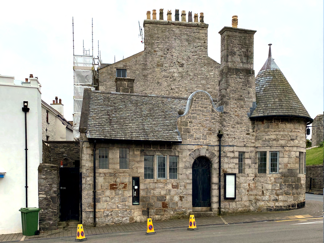 A photograph of a historic stone building featuring a turret and slate roof, with scaffolding visible on the left side indicating repair work.