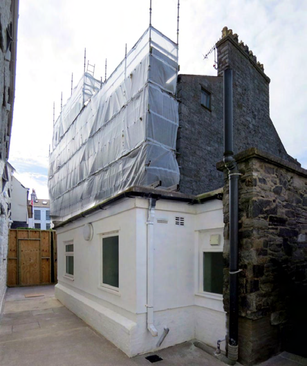 A photograph showing a building under renovation with scaffolding and sheeting covering the upper section, adjacent to a white single-story extension and a stone boundary wall.