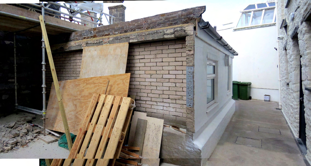 A photograph showing a construction site with scaffolding, exposed brickwork, and wooden boards, depicting the demolition or alteration of a rear extension.