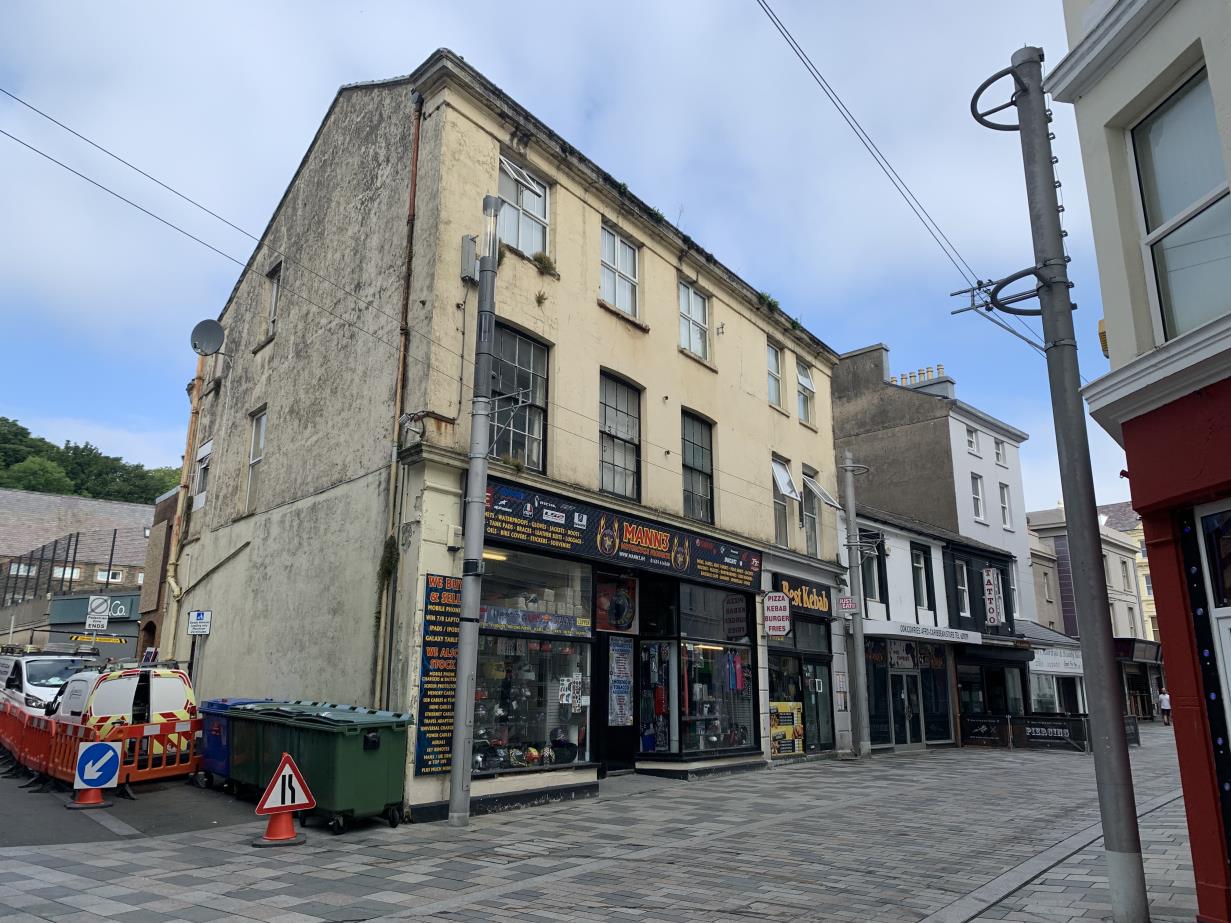 A street-level photograph of a weathered corner building featuring commercial shop fronts on the ground floor and windows above, with construction barriers and bins in the foreground.