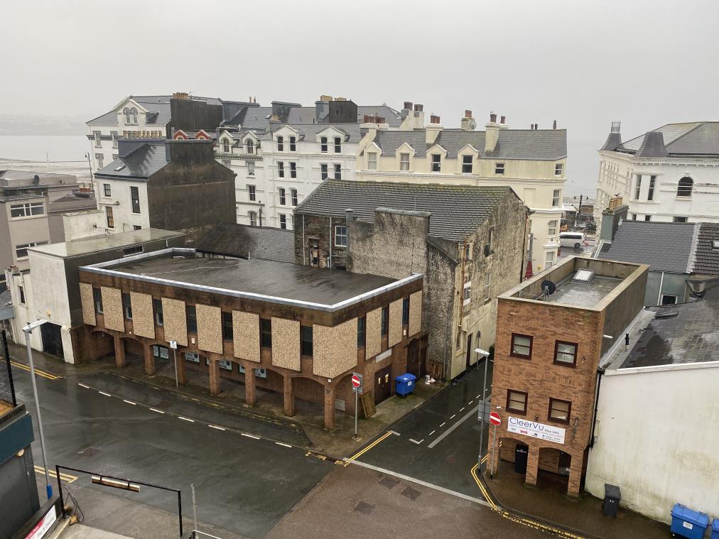 A high-angle photograph showing a street corner with a two-story commercial building and older stone structures, with the sea visible in the background.