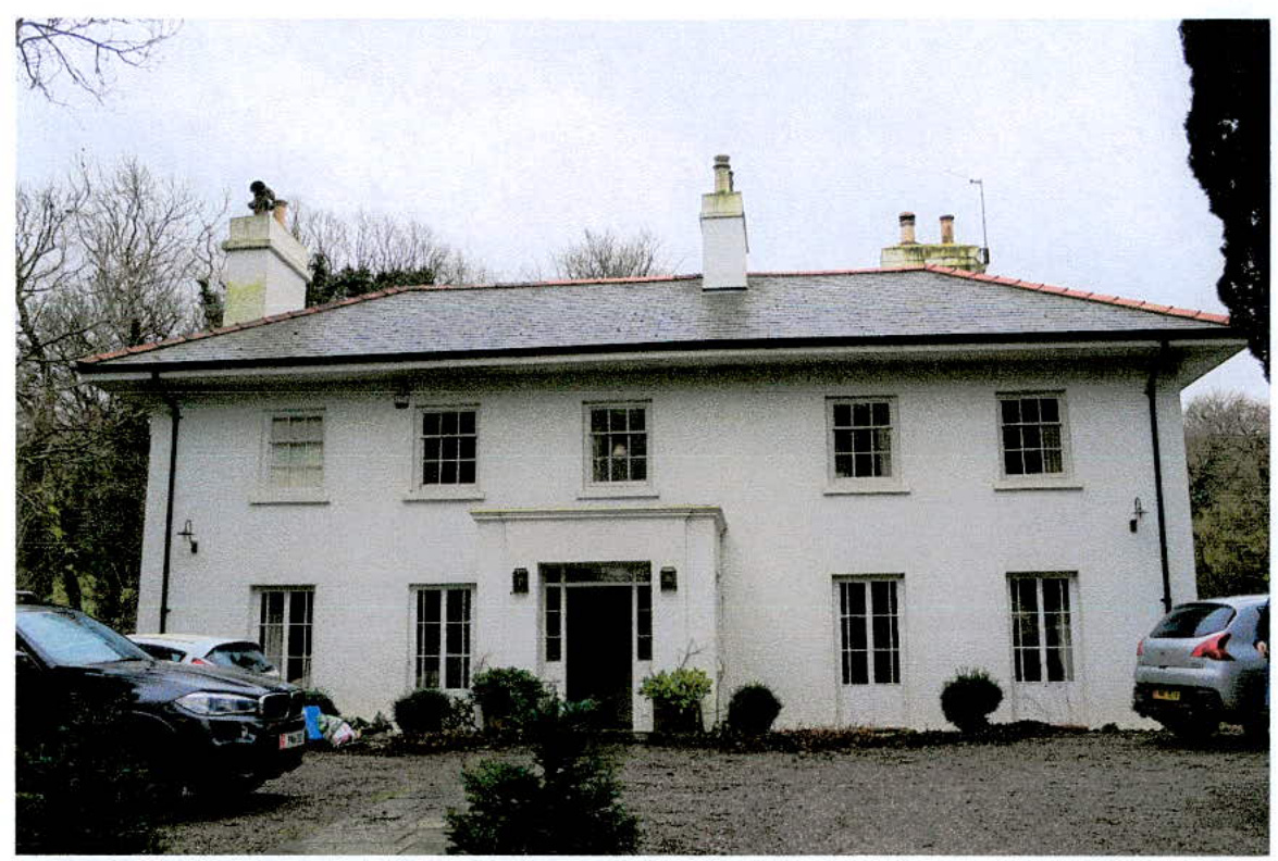 A photograph showing the front elevation of a large, white, two-story detached house with a slate roof and chimneys. Cars are parked on a gravel driveway in front of the property, which is surrounded by trees and shru...