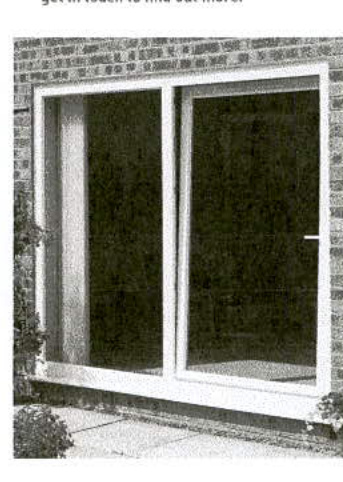 A black and white photograph showing a white uPVC window or patio door set into a brick wall with plants at the base.