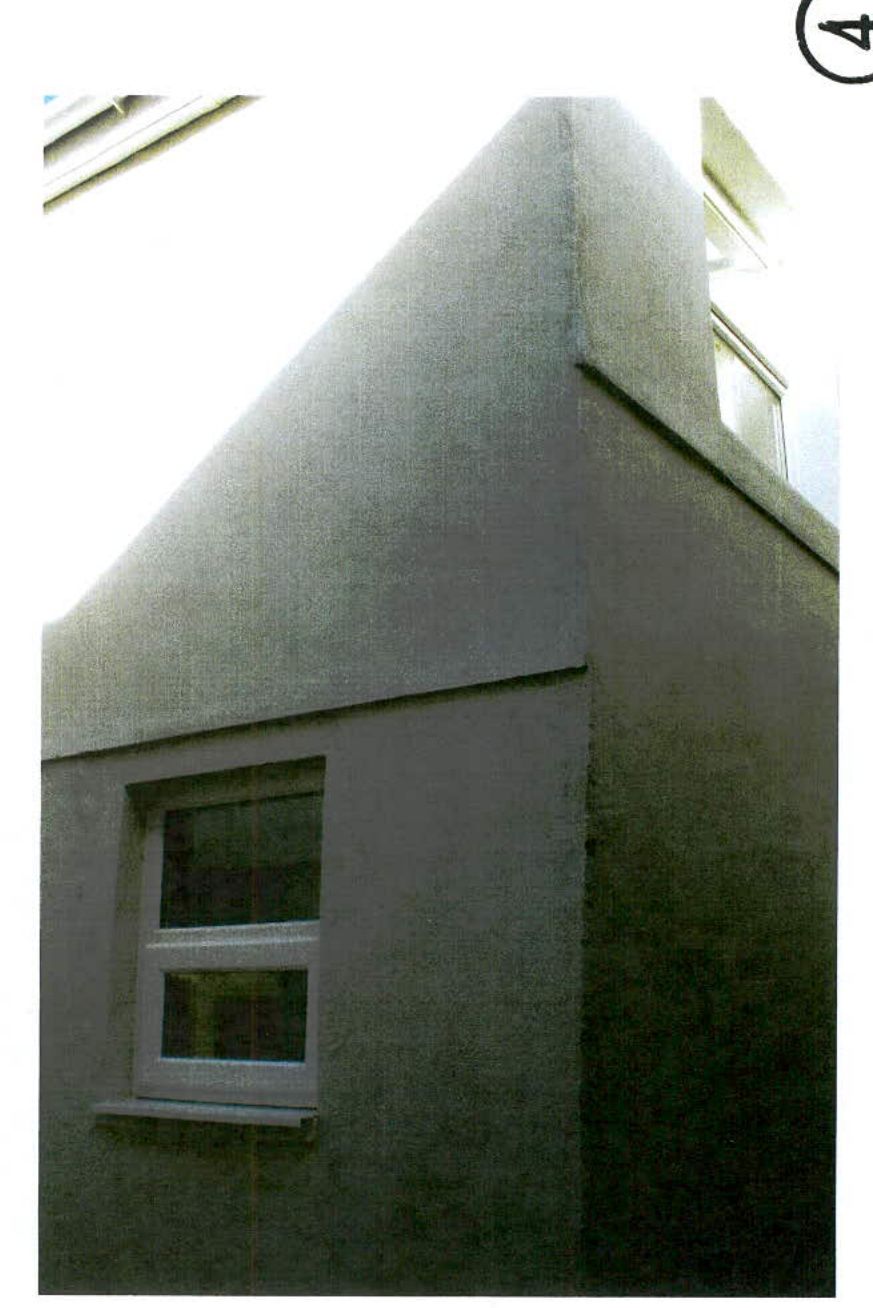 A close-up photograph showing the corner of a grey rendered building with a white window frame on the lower level and another window near the roofline.