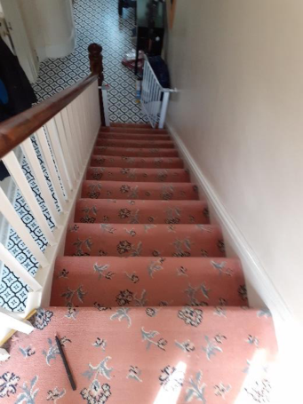 Interior photograph showing a residential staircase with pink floral carpet and a wooden banister. A white safety gate is installed at the top of the stairs near a tiled hallway floor.