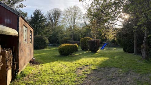 A photograph showing a grassy garden area with a wooden structure on the left, surrounded by trees and hedges.