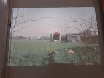A photograph of a rural site showing a grassy field, a small red shed, and an existing building to the right.