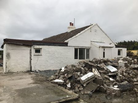 A photograph showing a white single-story bungalow with a large pile of rubble and construction debris in the foreground.