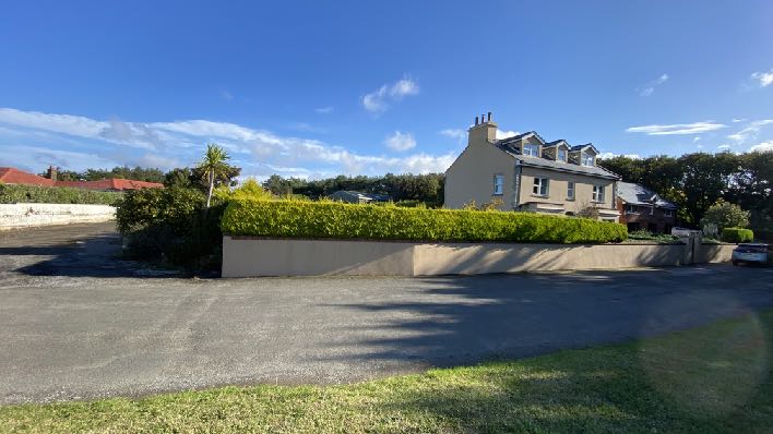 A street-level photograph showing a large detached house situated behind a retaining wall and hedge, with a paved road in the foreground.