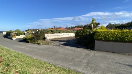 A photograph showing a paved access road or driveway leading towards a property with a stone boundary wall and houses in the background under a blue sky.