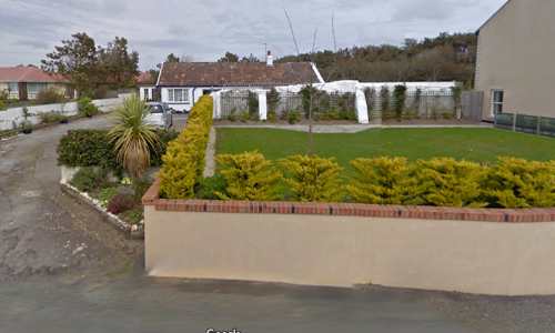 A photograph of a rural property featuring a white detached bungalow, a gravel driveway, and a large lawn bordered by a low wall and hedges.