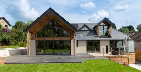 A modern single-story detached house featuring a prominent timber-clad gable end and large glass windows, photographed outdoors on a sunny day.