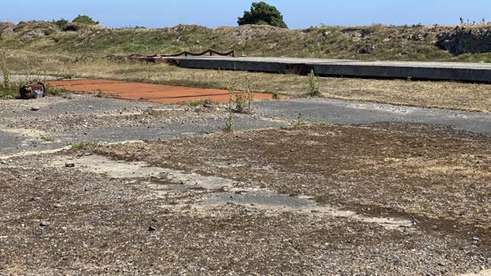 A photograph of a grassy, undeveloped plot of land featuring concrete slabs and a long concrete wall in the background, likely the site for the proposed nature reserve development.