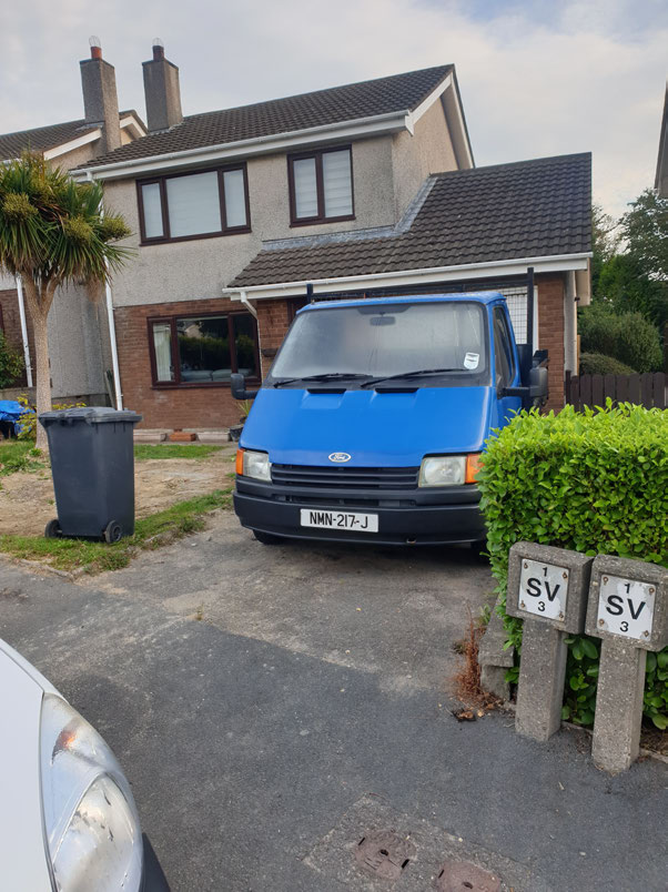 A photograph showing a blue van parked on a paved driveway in front of a two-story detached house with a palm tree and wheelie bin.